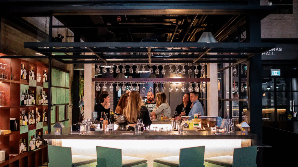 A group of people gathered around a modern bar at Waterworks Food Hall, talking and enjoying drinks while seated under hanging wine glasses, surrounded by shelves of bottles and warm ambient lighting.