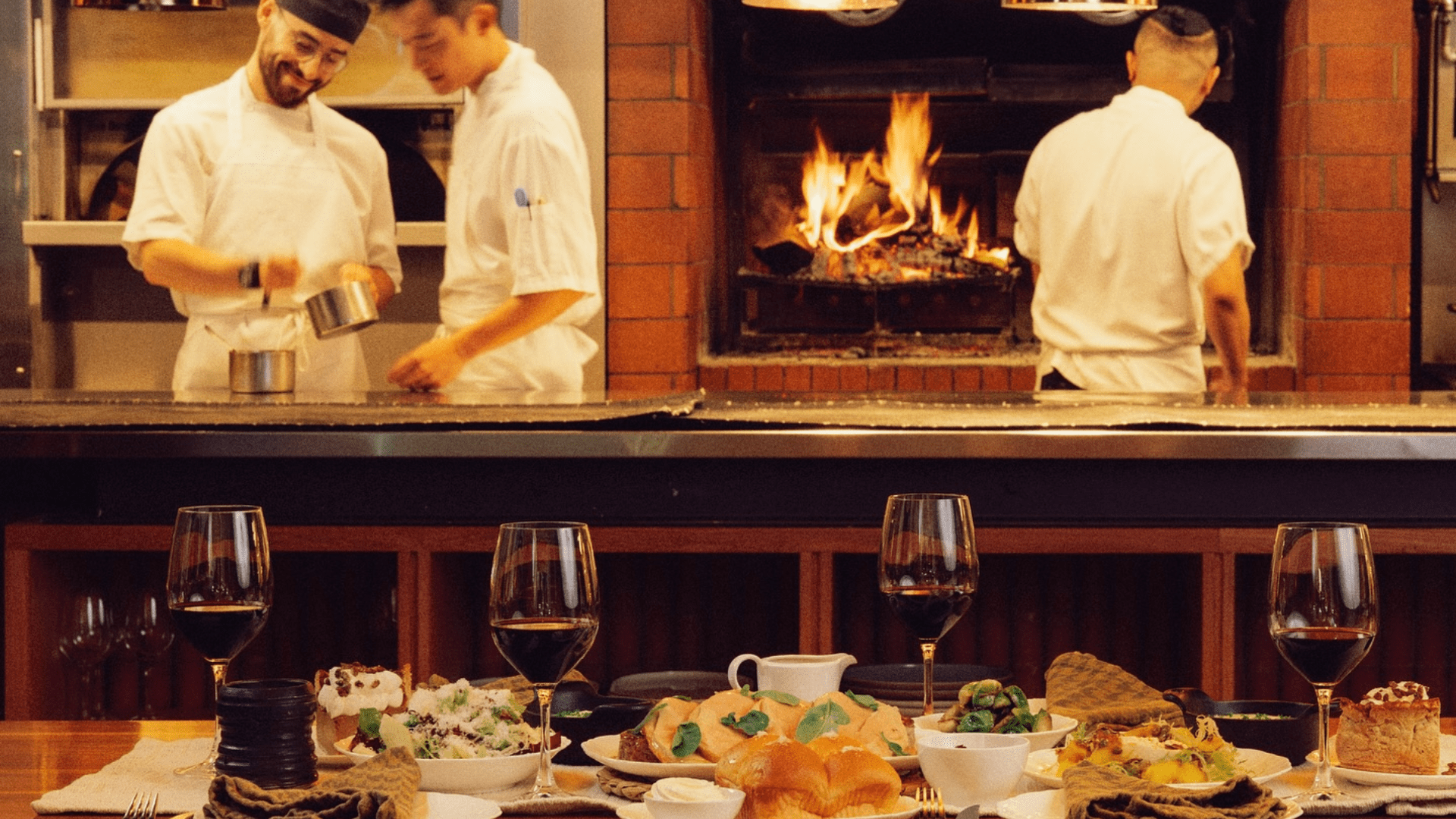 Open-kitchen restaurant scene with chefs cooking over a wood-fired hearth in the background and a foreground table set with wine glasses and plated dishes, showcasing an elevated dining experience.