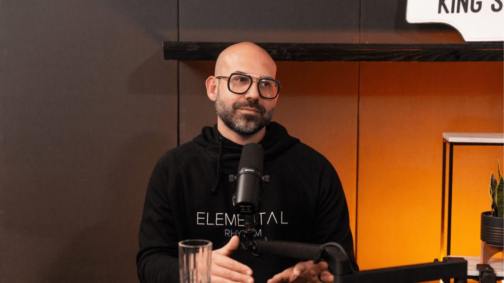 Gio Bartolomeo speaking into a microphone during a Table 6ix podcast recording, seated in a warm, softly lit studio.