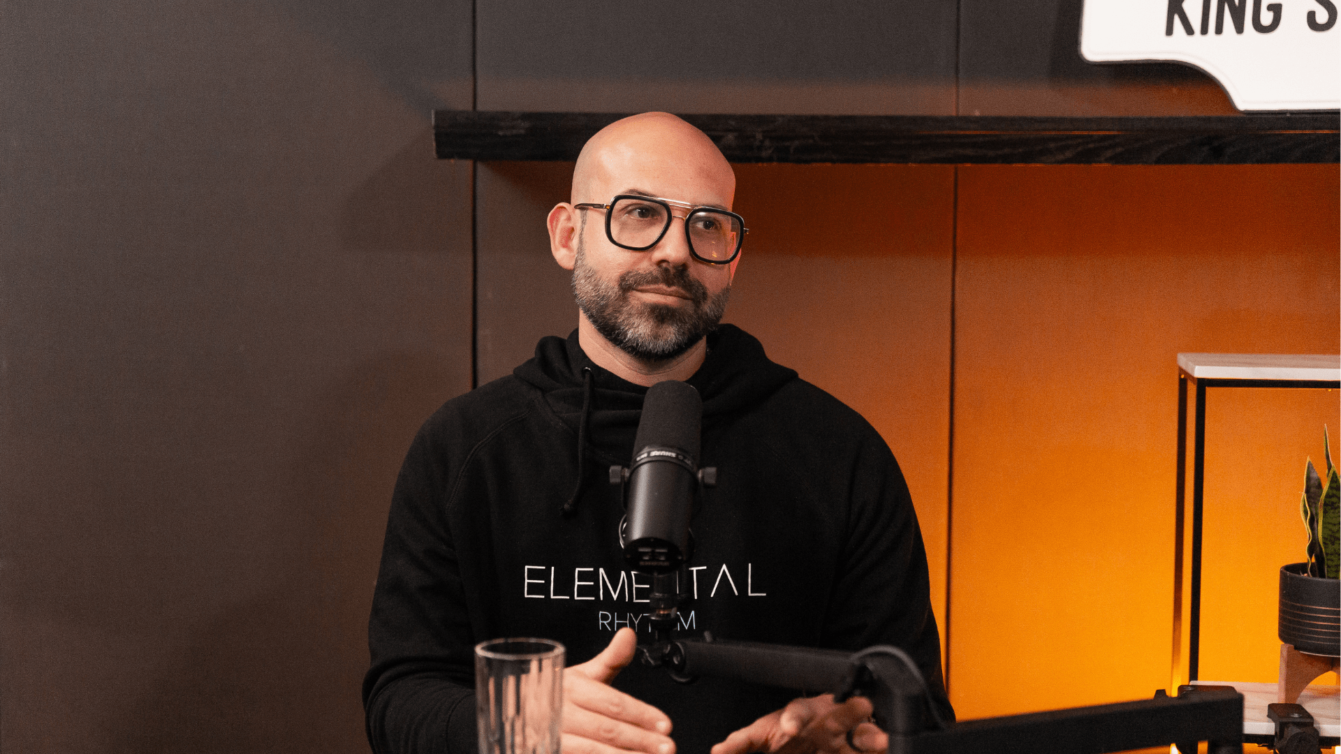 Gio Bartolomeo speaking into a microphone during a Table 6ix podcast recording, seated in a warm, softly lit studio.