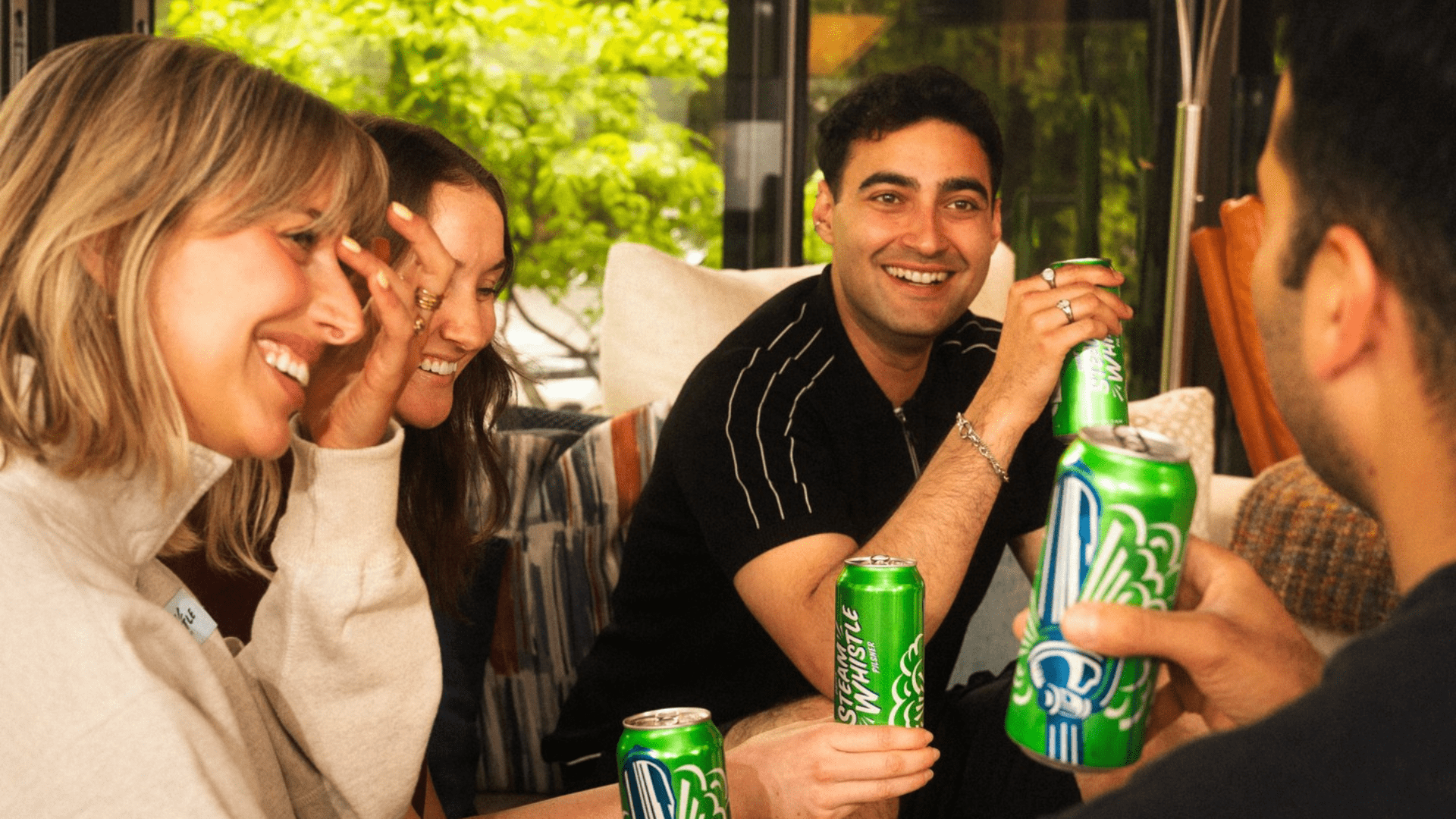 A group of adults smiling and chatting while holding green beer cans, seated together indoors at a social gathering with natural light and greenery visible in the background.