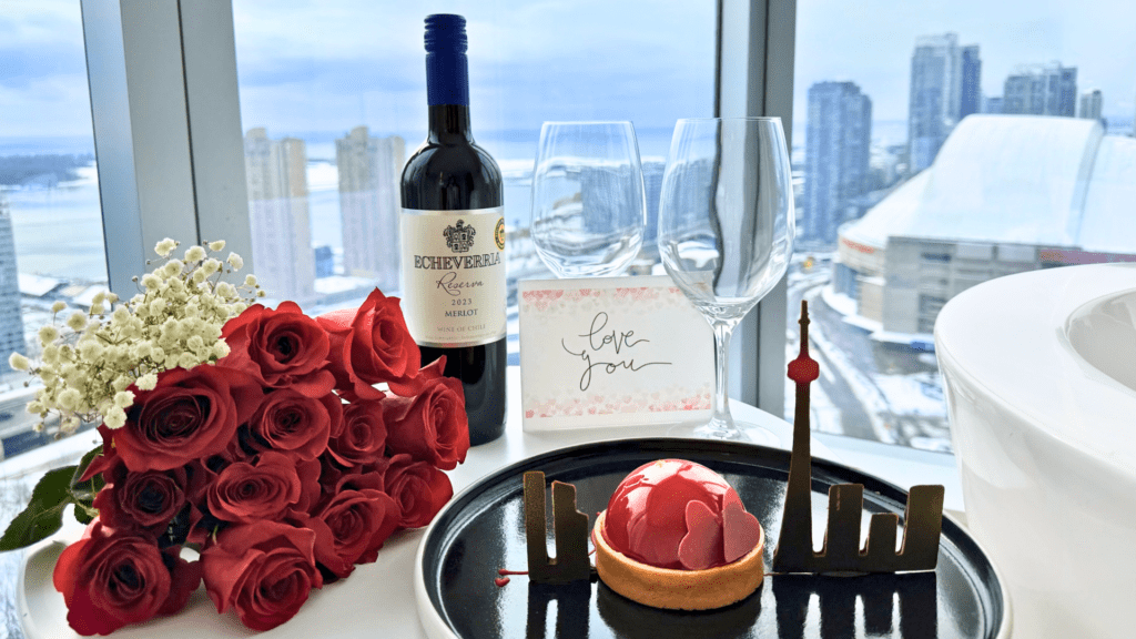Valentine’s Day setup in a downtown Toronto hotel room featuring a bouquet of red roses, a bottle of red wine, two wine glasses, a handwritten “love you” card, and a heart-shaped dessert with a chocolate CN Tower silhouette, set against skyline views through floor-to-ceiling windows.
