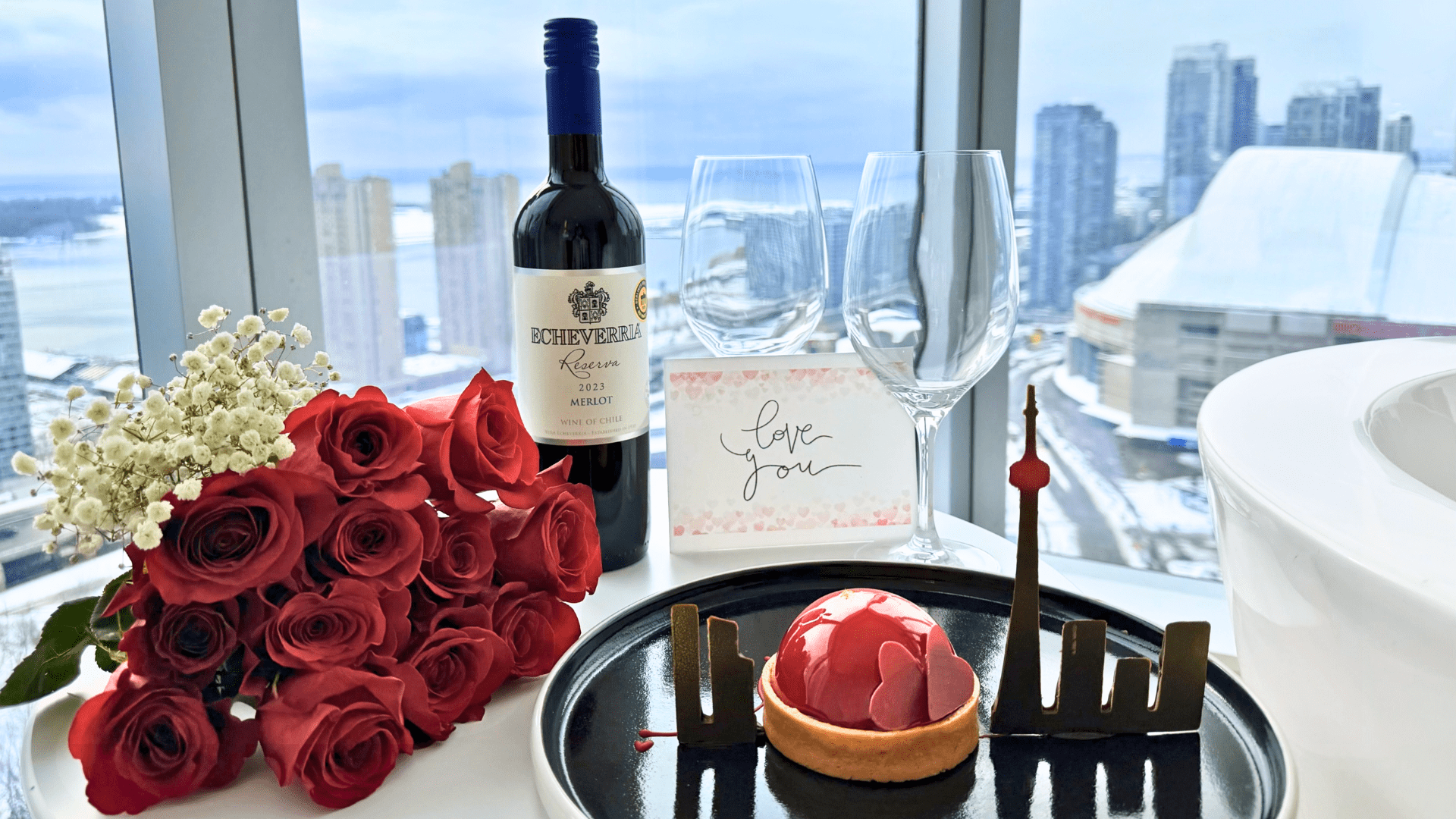 Valentine’s Day setup in a downtown Toronto hotel room featuring a bouquet of red roses, a bottle of red wine, two wine glasses, a handwritten “love you” card, and a heart-shaped dessert with a chocolate CN Tower silhouette, set against skyline views through floor-to-ceiling windows.