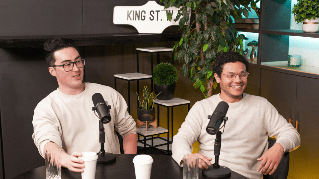 Aki Erenberg and Ricky Barrientos sit at a podcast table with microphones, smiling during a Table 6ix recording, with a King St. sign and greenery in the background.