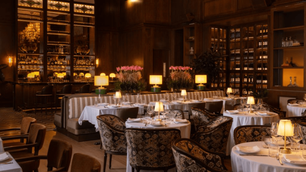 Elegant restaurant interior with warm wood-paneled walls, softly lit table lamps, white tablecloth dining tables, patterned upholstered chairs, and a glowing back bar lined with bottles and floral arrangements, creating an intimate, upscale atmosphere.