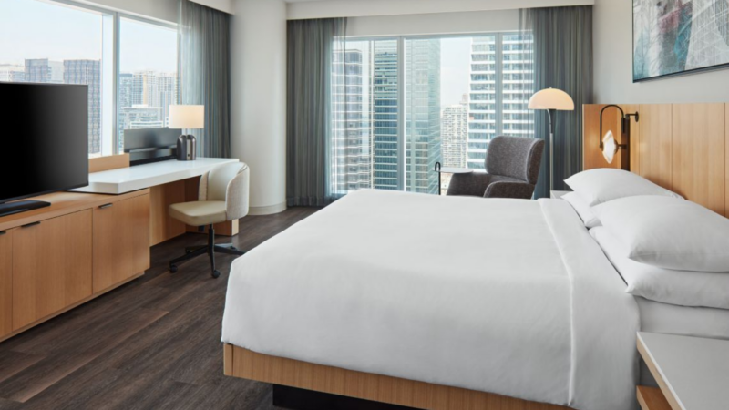 Modern hotel room in Downtown Toronto with a king bed, large floor-to-ceiling windows overlooking the city skyline, a workspace desk, and minimalist contemporary furnishings.