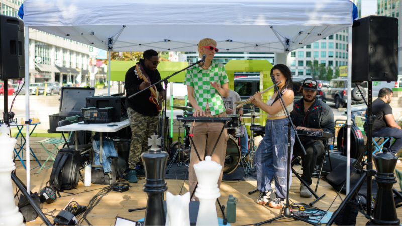 Live band performing at an outdoor setup under a white canopy at The Deck in Clarence Square, with musicians playing keyboard, guitar, flute, and drums. Large chess pieces are visible in the foreground, with city buildings and a street scene in the background.