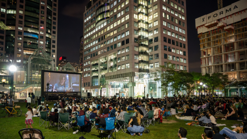 Crowds watching an outdoor movie screening at David Pecaut Square in downtown Toronto at night.
