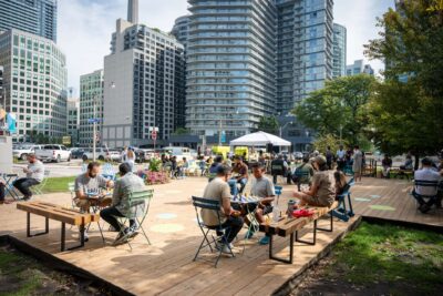 Groups of people play chess on an outdoor urban wooden deck, nestled between condominiums and trees in downtown Toronto.